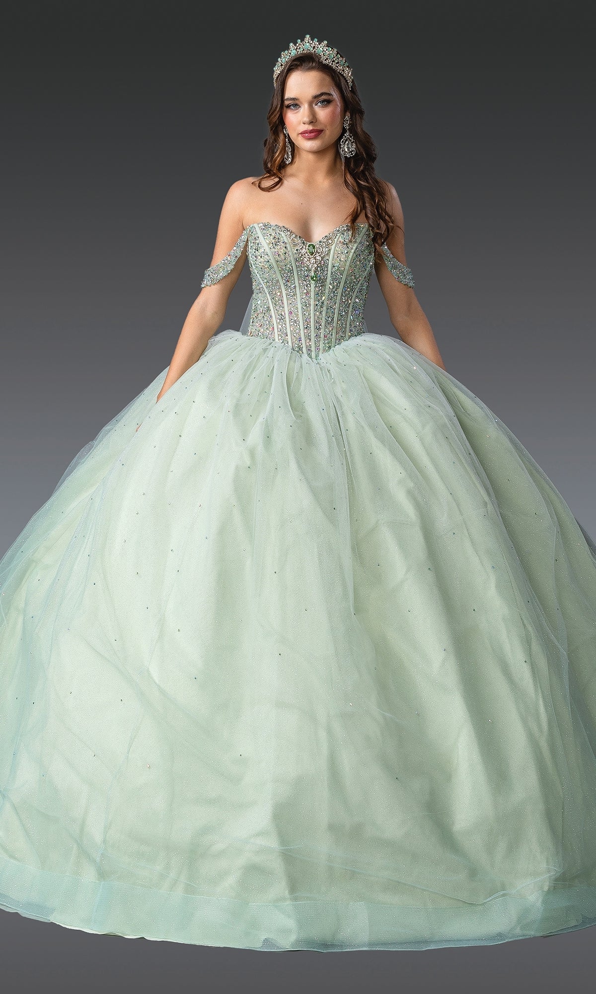 A woman in the Light Green Quinceanera Dress 1950 By Dancing Queen, featuring a beaded corset bodice and glitter tulle skirt with matching arm bands, tiara, and earrings, stands against a neutral background.